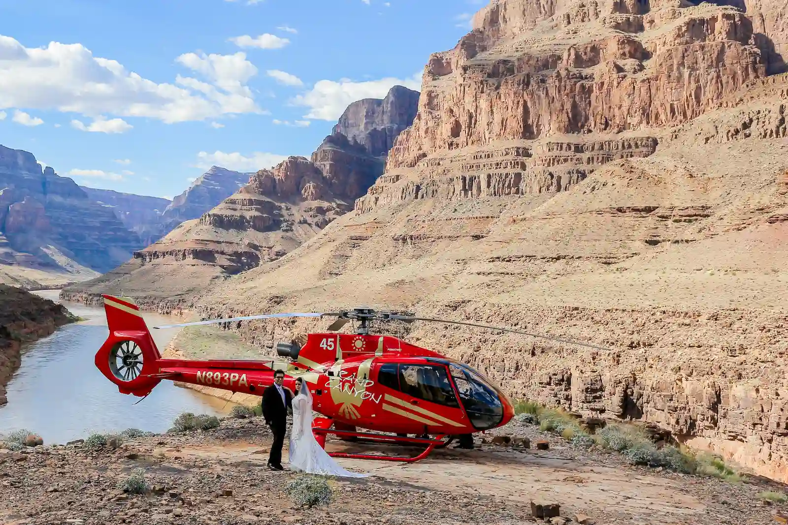Bride and groom posing beside a red Grand Canyon helicopter at the bottom of the canyon with the Colorado River in the background
