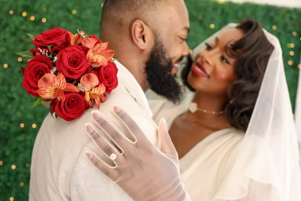 Bride and groom embracing in front of a greenery wall at a Las Vegas wedding, bride holding red rose bouquet with orange flowers